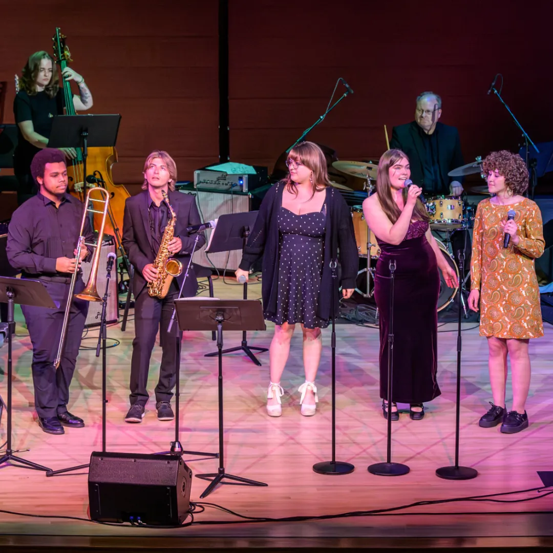 Six musicians perform on a stage with microphones, including a trombonist, a saxophonist, three vocalists, and a drummer in the background.