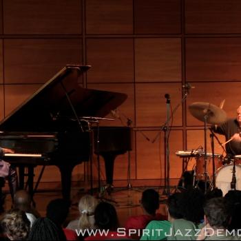 A pianist in a purple jacket and a drummer perform on stage in front of a seated audience; microphones and instruments are visible.