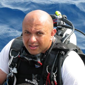 A person in scuba diving gear surfaces from the ocean, wearing a wetsuit and dive equipment against a background of blue water.