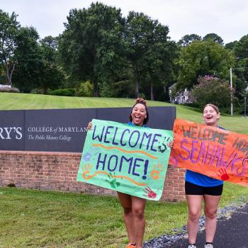 Two smcm Orientation Leaders hold signs that read "Welcome Home" and "Welcome Home Seahawks" in front of the College Drive on new student move-in day. 
