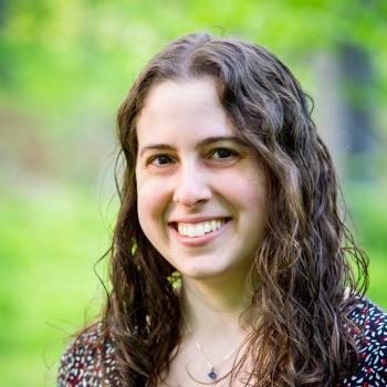Dr. Gili Freedman, person with long brown curly harir smiling in front of a blurred image of greenery
