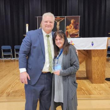 A man in a suit stands next to a woman holding an award on a wooden stage with a religious backdrop featuring a crucifixion image and candles. Empty chairs are arranged behind them.
