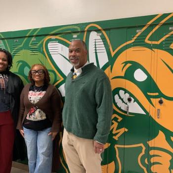 Three people stand smiling in front of green lockers with a large yellow and green bee mascot painted on them. The two women are on the left, one in a graphic T-shirt and blazer, the other in glasses and a cardigan. The man on the right wears a green sweater.