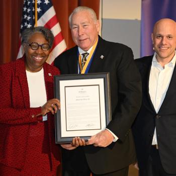 Three people stand together, smiling. The woman on the left is holding a framed document with the man in the center. An American flag and a blue banner are in the background. The man on the right is wearing a suit and has a pin on his lapel.