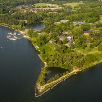 Aerial view of a picturesque landscape featuring a lush, green shoreline surrounded by water. The area includes buildings, roads, and a marina with boats, all encompassed by dense tree cover and grassy fields.