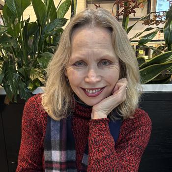 A woman with shoulder-length blonde hair and a red patterned sweater smiles, resting her chin on her hand. 