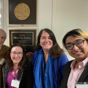 Four people are smiling and standing in front of a wall plaque for Senator Chris Van Hollen, Maryland. The group includes one person with glasses and another wearing a blue scarf. They appear to be in an office setting.