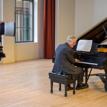A person wearing a gray suit plays a grand piano in a spacious, well-lit room with large windows and beige walls. A video camera on a tripod is partially visible on the left, recording the performance.