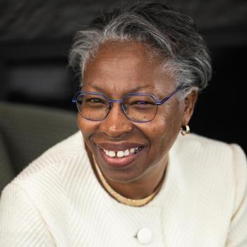 Smiling woman wearing glasses and a white textured blazer, sitting in front of a dark background. Her hair is short and gray, and she is accessorized with small gold hoop earrings.