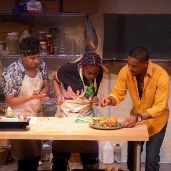 Three people stand at a kitchen counter with food. One person in a yellow shirt points at a dish, while two others wearing aprons watch intently. Shelves with cooking supplies are in the background.