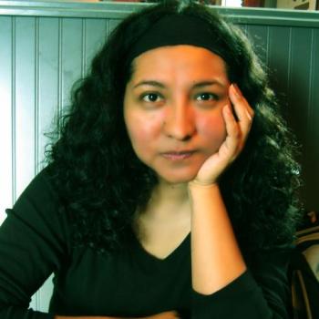 Person with long curly hair and a headband, resting their chin on their hand. They're seated indoors against a striped wall, wearing a dark top and looking toward the camera.