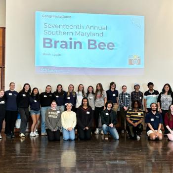 A group of 28 people stand and kneel in rows on a stage, smiling, in front of a large screen displaying text: "Seventeenth Annual Southern Maryland Brain Bee, March 1, 2025