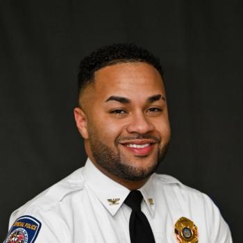 A man in a white police uniform with badges and a name tag that reads "Martinez" smiles against a dark backdrop.