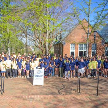 A large group of people standing outside in front of a brick building, gathered around a "Volunteer Check-In" signpost on a sunny day.