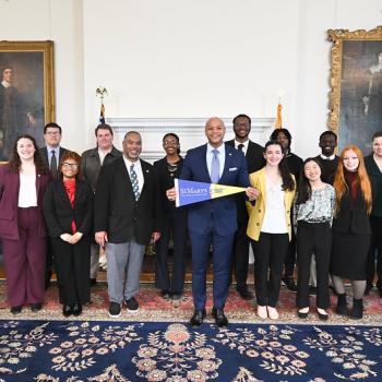 A group of people, dressed in business attire, stand together in a formal room with paintings on the walls. One person in the center holds a St. Mary's sign.