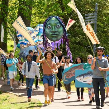 A group of people march outdoors holding banners, flags, a large puppet head, and a cardboard fish, with trees and greenery in the background.
