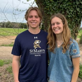 Two people standing outdoors, smiling at the camera. One wears a St. Mary's College rowing shirt. Green field and tree in the background.