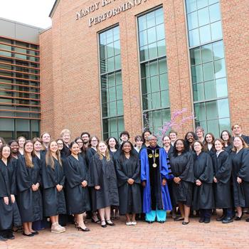 Group of graduates in black gowns posing with faculty member in blue regalia outside the Dodge Performing Arts Center. One holds a large key-shaped sign.