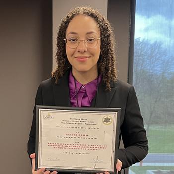 A person wearing glasses and a suit stands indoors holding a framed certificate and smiling at the camera.