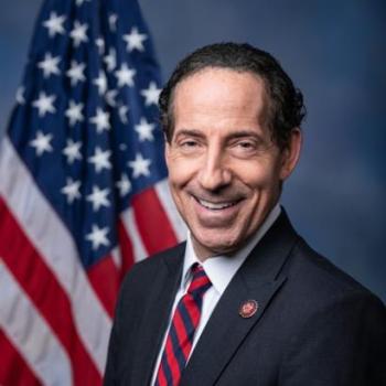 A man in a dark suit and striped tie stands in front of a U.S. flag, smiling at the camera against a blue background.