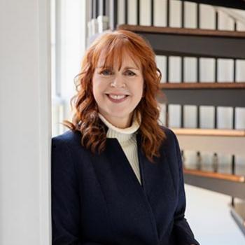 A woman with red hair, wearing a dark blazer and light turtleneck, stands indoors near a window, smiling with hands clasped. A spiral staircase is visible in the background.