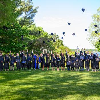 A group of graduates in caps and gowns stand on a lawn, tossing their caps into the air in celebration, surrounded by green trees under a clear sky.