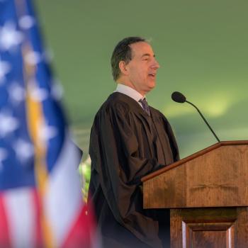 A man in academic robes speaks at a podium labeled "St. Mary’s College" during an outdoor event, with an American flag visible in the foreground.
