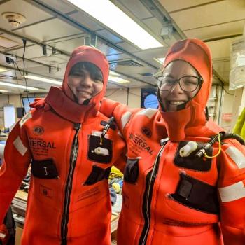 Two people wearing orange immersion survival suits smile inside a room filled with equipment and supplies.
