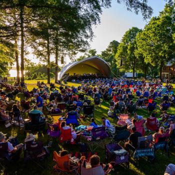 Large crowd sitting on lawn chairs and blankets at an outdoor concert in a park, with a stage set up under trees on a sunny day.