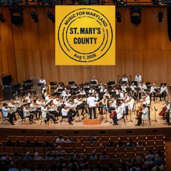 A symphony orchestra performs on stage in a wooden concert hall with a banner overhead reading "Music for Maryland, St. Mary's County, Aug. 1, 2025." Audience members are seated below.