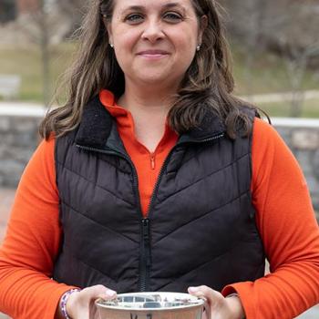 A woman in an orange shirt and black vest stands outdoors, holding a silver bowl with both hands and looking at the camera.