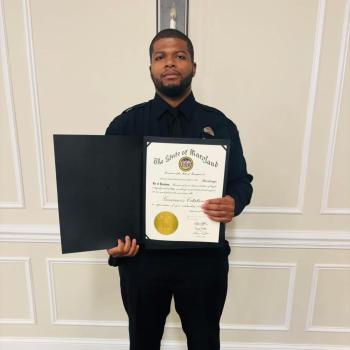 A man in a dark uniform holds an open certificate folder displaying an official document from the State of Maryland, standing in front of a beige paneled wall.