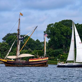 A wooden tall ship with a race committee flag and a modern white sailboat are side by side on a river, with trees and greenery in the background.