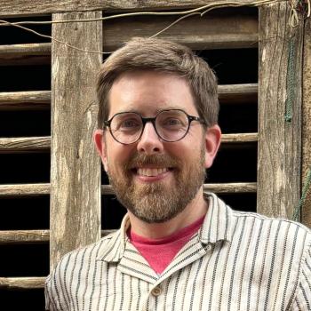 A bearded man with glasses and short hair, wearing a striped shirt and red t-shirt, stands in front of a weathered wooden window.