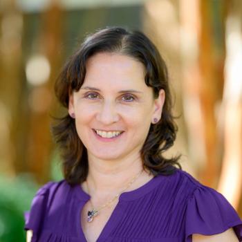 A woman with shoulder-length brown hair and a purple blouse smiles at the camera, standing outdoors with a blurred background.