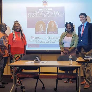 Four people stand in front of a projector screen displaying information about a community conversation event at Montgomery College. A table with chairs is in the foreground.