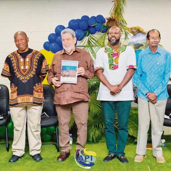Six men stand side by side indoors; one holds a book or magazine. There are balloons, plants, and a decorated wall behind them, with an "API" logo on the grass at their feet.