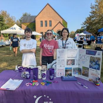 Three people stand behind a Relay for Life booth at an outdoor event, with informational materials and promotional items displayed on the table. Tents and people are visible in the background.