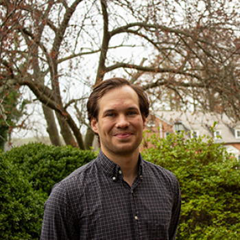 A man stands outdoors in front of green bushes and leafless trees, wearing a checked button-up shirt, smiling at the camera.