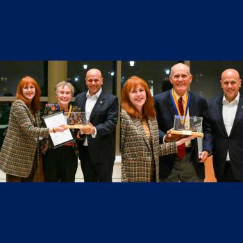 Six people stand indoors, with two women and one man holding awards and certificates, all dressed in formal or business attire, smiling at the camera.