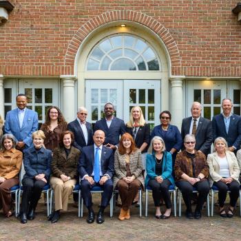 A group of 24 adults pose for a formal group photo in two rows outside a brick building with arched windows and columns.