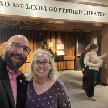 Two people smiling for a selfie inside a theater lobby with a sign reading “Brad and Linda Gottfried Theater” above them; several others are in the background.