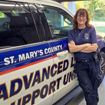A person in a navy uniform stands smiling and leaning against a St. Mary's County Advanced Life Support Unit vehicle, parked inside a garage.