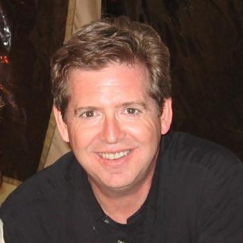 A man with short brown hair and a black shirt smiles at the camera in an indoor setting.