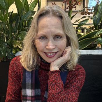 A woman with blonde hair wearing a red patterned sweater and plaid scarf sits indoors, smiling with her chin resting on her hand. Green plants and a dark partition are in the background.