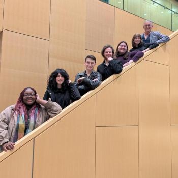 Seven people pose on a staircase inside a building with tan walls; six stand on the steps while one stands at the base, all facing the camera.
