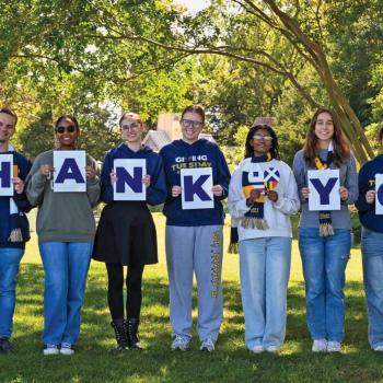 Eight people stand outdoors in a row, each holding a letter sign that together spell out "THANK YOU," with trees and greenery in the background.