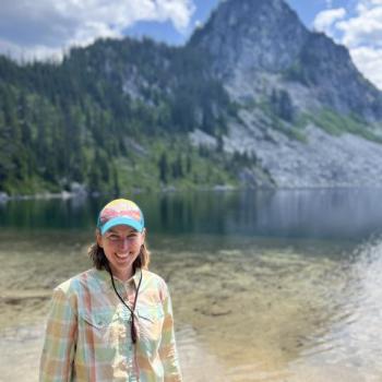 A person wearing a plaid shirt and colorful cap stands near a clear mountain lake with a forested, rocky peak in the background.