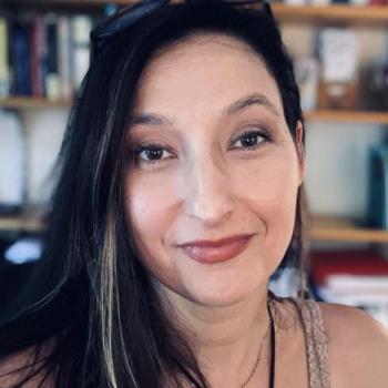 A woman with long brown hair and light makeup smiles softly at the camera, with bookshelves in the blurred background.