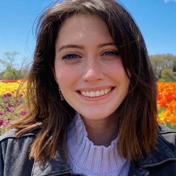 A woman with shoulder-length brown hair smiles outdoors in front of colorful flowers under a blue sky.